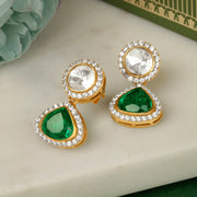 A pair of silver earrings with green stones and white accent stones, displayed on a marble surface with a green fabric backdrop.